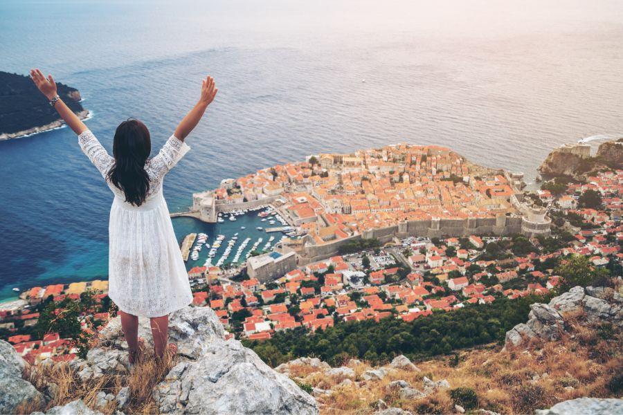A woman facing away from the camera is standing on a cliff with her hands in the air and taking in the panorama of Dubrovnik below