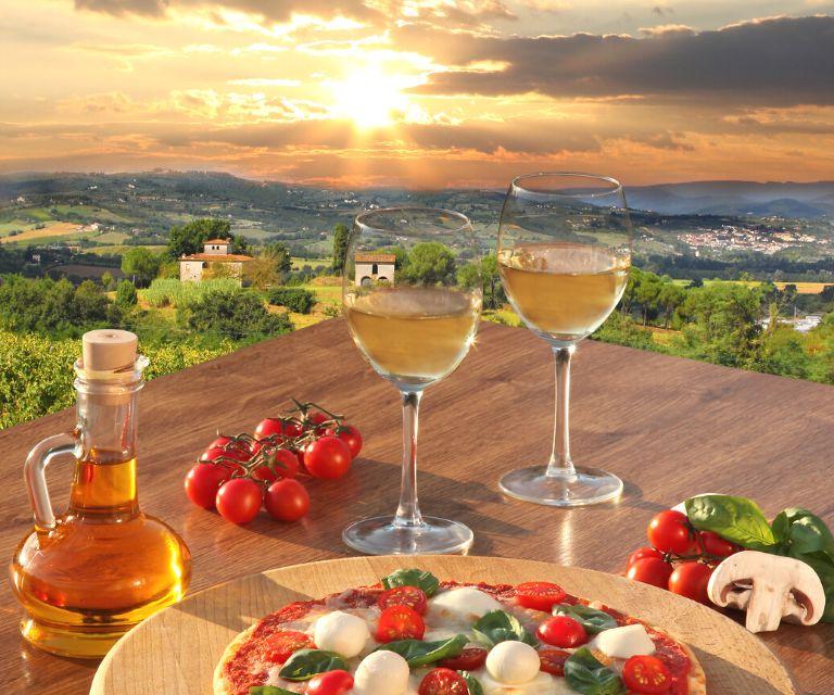 two glasses of white wine on a table, there is also a bottle of olive oil, pizza, and some vegetables.