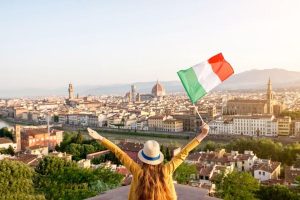 A woman in a sunhat and yellow top is facing away from the camera. She helds her arms in the air and holds an Italian flag in her righ hand, clearly enjoing her city break to Florence
