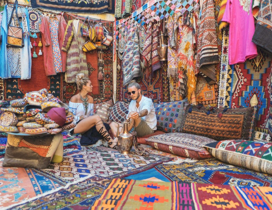 Man and a woman are sitting among various beautiful carpets as they visit a market stall that sells them.